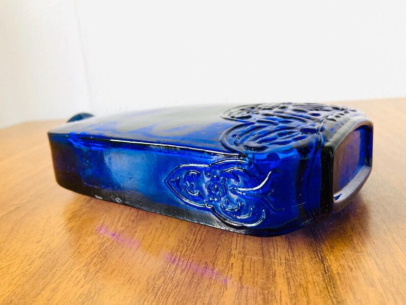 May include: A rectangular cobalt blue glass bottle with ornate embossed floral and scroll designs. The bottle has a rounded top and is resting on a wooden surface.