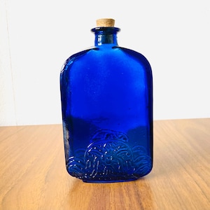 Puede incluir: Una botella de vidrio azul cobalto con tapón de corcho. La botella tiene forma rectangular con bordes redondeados y un diseño decorativo en relieve cerca de la base. Se encuentra sobre una superficie de madera.