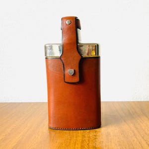 Può includere: Una fiaschetta rettangolare in pelle marrone con la parte superiore argentata e un cinturino in pelle. La fiaschetta ha un piccolo stemma sul davanti e si trova su una superficie di legno.