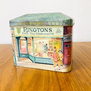 Peut inclure: Une boîte à thé en métal rectangulaire avec un toit vert. La face avant présente une illustration d'une devanture de magasin marquée Ringtons Fine Tea Merchants Est. 1907, avec deux personnages devant et un commerçant à l'intérieur.