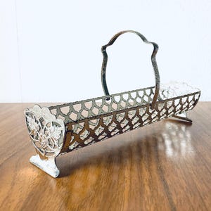 May include: An antique silver-coloured metal basket with a handle. The basket has a rectangular shape with a decorative openwork design. The ends of the basket feature ornate floral and leaf patterns. The handle is arched and the basket sits on small feet.