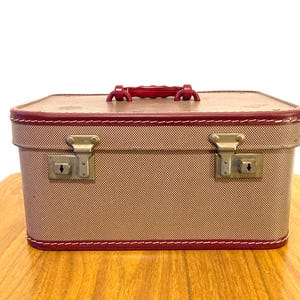 Pode incluir: Uma mala vintage com exterior bege e acabamento vermelho. A mala tem uma alça vermelha e dois fechos de metal. Os cantos são reforçados com acabamento vermelho e costuras brancas. A mala está sobre uma superfície de madeira.