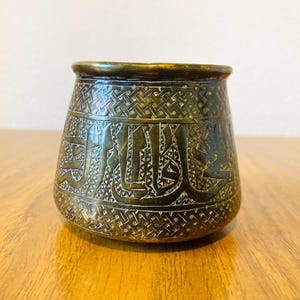 May include: A small, antique bronze-coloured pot with intricate etched patterns and Arabic script. The pot has a wide mouth and a slightly rounded base. The surface is detailed with geometric designs and calligraphy.