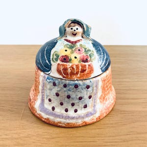 May include: A ceramic trinket box in the shape of a woman, with a blue shawl and bonnet. The lid depicts a basket of colourful flowers. The base is orange with a blue apron and purple polka dots.