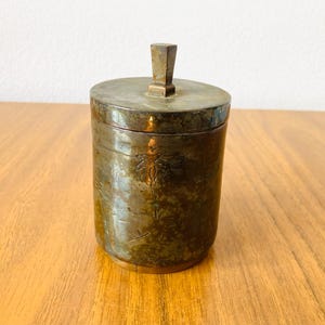Könnte beinhalten: Ein zylindrischer antiker Messingbehälter mit Deckel und quadratischem Griff. Der Behälter hat geätzte Blumenmuster und steht auf einer Holzoberfläche. Das Messing hat ein angelaufenes, gealtertes Aussehen.