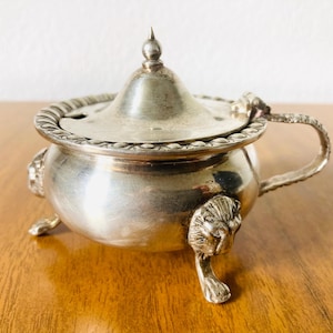 May include: An antique silver-coloured sugar bowl with a lid and handle. The bowl has a round body, rests on three lion's paw feet, and features a decorative rim. The lid is topped with a small finial.