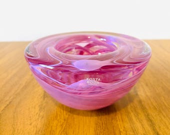 May include: A round, pink glass bowl with a swirling design. The bowl has a wide opening and a rounded base. The glass is translucent, allowing light to pass through. The brand name "KOSTA BODA" is visible on the side.