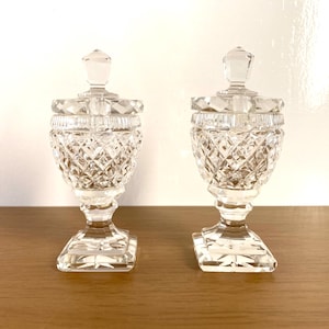 Puede incluir: Dos tarros de cristal transparente con tapas. Los tarros tienen un patrón de diamantes y se asientan sobre una base cuadrada. Las tapas están rematadas con un remate de cristal. Los tarros se exhiben sobre una superficie de madera.