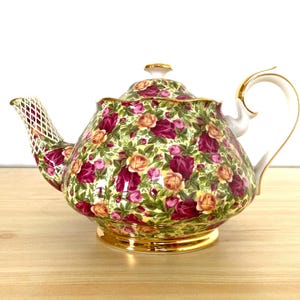Könnte beinhalten: Eine Teekanne mit floralem Muster und goldenen Verzierungen und Griff. Die Teekanne hat ein Design aus roten und gelben Rosen auf grünem Hintergrund. Der Ausguss hat ein Gittermuster.