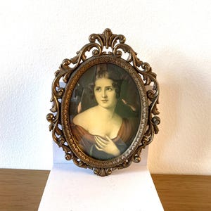 Puede incluir: Retrato ovalado antiguo en un marco decorativo de bronce. El retrato presenta a una mujer con un vestido burdeos. El marco tiene un diseño ornamental y con volutas. El retrato se muestra sobre una superficie blanca.