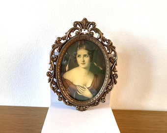 Puede incluir: Retrato ovalado antiguo en un marco decorativo de bronce. El retrato presenta a una mujer con un vestido burdeos. El marco tiene un diseño ornamental y con volutas. El retrato se muestra sobre una superficie blanca.