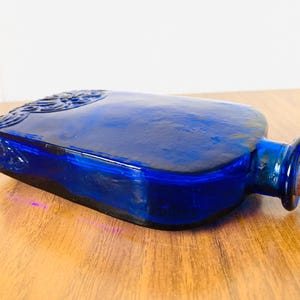 May include: A cobalt blue glass bottle with a rectangular shape and a cork stopper. The bottle has embossed detailing on the top and sides. The bottle is lying on a wooden surface.