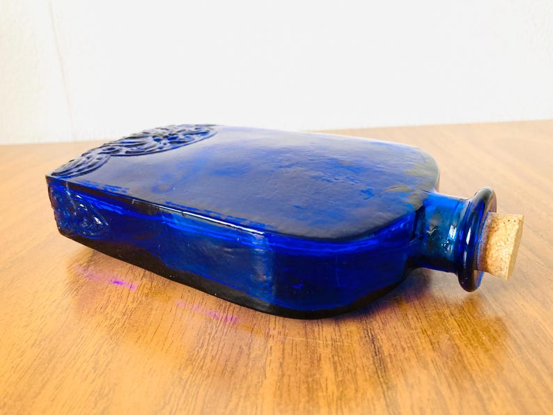 May include: A cobalt blue glass bottle with a rectangular shape and a cork stopper. The bottle has embossed detailing on the top and sides. The bottle is lying on a wooden surface.
