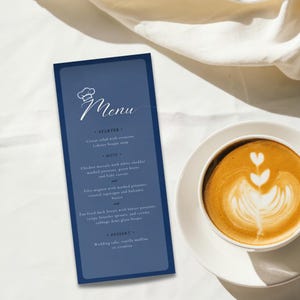 May include: A blue menu card with a white chef hat icon and the word "Menu" in white cursive script. The menu lists a starter, main course, and dessert options. The text includes "Caesar salad with croutons", "Lobster bisque soup", "Chicken marsala", "Filet mignon", "Pan fried duck breast", "Wedding cake", "vanilla muffins", and "tiramisu".