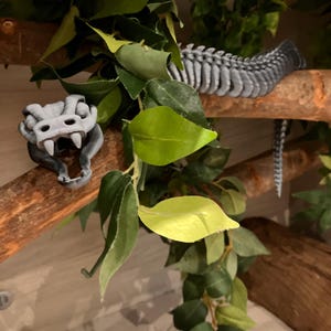 May include: A grey plastic snake skeleton hanging from a branch with green leaves. The snake skeleton is made of individual bones connected together.