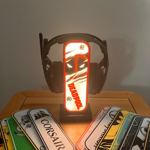 May include: A black headphone stand with a light-up Deadpool logo. The stand is surrounded by various colourful stickers with logos like Corsair, Google, and a bear.
