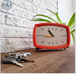 May include: A vintage-style red clock with a cream face and black markings. The clock has a grid pattern and green legs. A set of keys lies on a wooden surface in the foreground. The clock's dimensions are approximately 15 cm in width.