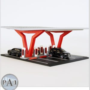 May include: A miniature electric vehicle charging station model featuring a white roof supported by red angular pillars. The base is a black parking lot with white painted lines, and it includes several small toy cars parked at white charging units.