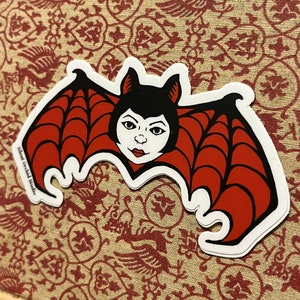 May include: A red and black bat sticker with a white outline. The bat has a webbed wing design and a cartoon face with black hair and red lips. The sticker has the text "Silent Orchid Studio" in the bottom left corner.