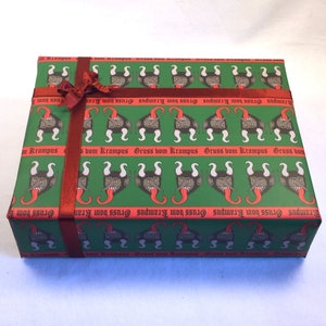 May include: A green gift wrap with a repeating pattern of a black and red devil's head with horns and a tongue sticking out. The text "Gruss vom Krampus" is repeated in red on the wrapping paper. A red ribbon is tied around the box.
