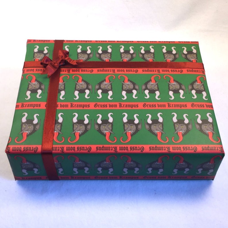 May include: A green gift wrap with a repeating pattern of a black and red devil's head with horns and a tongue sticking out. The text "Gruss vom Krampus" is repeated in red on the wrapping paper. A red ribbon is tied around the box.
