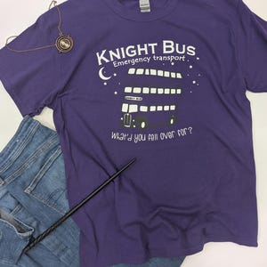 May include: Purple t-shirt with a white graphic of the Knight Bus from the Harry Potter series. The graphic includes the text "Knight Bus", "Emergency transport", and "What'd you fall over for?"