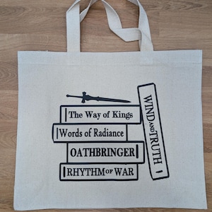 May include: A white canvas tote bag with a black design of a stack of books with the titles "The Way of Kings", "Words of Radiance", "Oathbringer", and "Rhythm of War". The design also includes a sword and the words "Wind and Truth" on the side of the stack of books.
