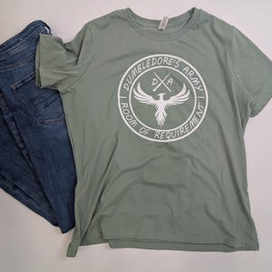 May include: A sage green t-shirt with a white graphic of a phoenix with wings spread. The graphic is inside a circle with the text "Dumbledore's Army Room of Requirement" around the outside.