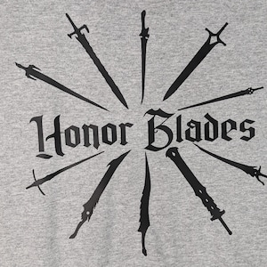 May include: A gray t-shirt with a black graphic design of swords radiating out from the center. The text "Honor Blades" is in a stylized font.