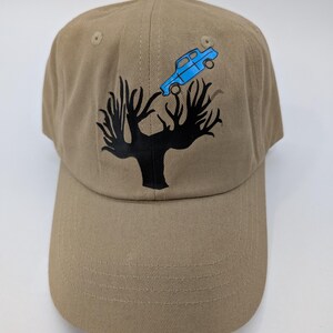 May include: A tan baseball cap with a black tree graphic and a blue car graphic. The cap has a curved brim and an adjustable strap at the back. The cap is made of a durable fabric.