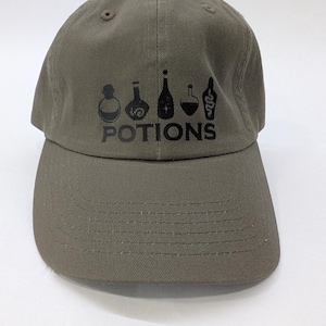 Può includere: Cappellino da baseball verde oliva con la scritta "POTIONS" in nero, accompagnata da illustrazioni nere di bottiglie di pozioni. Il cappellino ha una visiera curva e un cinturino in tessuto regolabile.