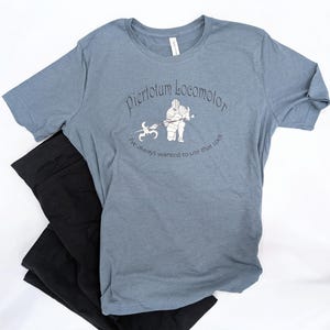 Op de afbeelding: Een blauw t-shirt met de tekst "Piertotum Locomotor" en een afbeelding van een ridder. De tekst "I've always wanted to use that spell" staat eronder. Een zwarte broek is onder het shirt gevouwen.