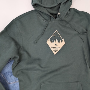 May include: A dark green hooded sweatshirt with a pocket on the front. The sweatshirt has a beige diamond-shaped graphic with a crescent moon, stars, trees, and a wolf silhouette. The graphic says "Forbidden Forest National Park".