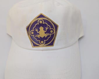 May include: White baseball cap featuring a purple and gold emblem with a frog design. The cap has a curved brim and a classic six-panel construction. The emblem is centered on the front of the cap.