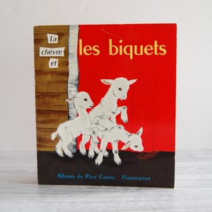 May include: Vintage children's book titled "La chèvre et les biquets." The cover features a red background with white illustrations of three goats. The book's spine is brown and white, with text in French. Published by Albums du Père Castor Flammarion.