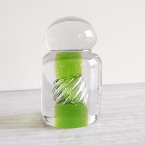 May include: A clear glass decorative bottle with a rounded top and a green interior. The bottle has a spiral design in the center. The bottle is sitting on a light gray surface.