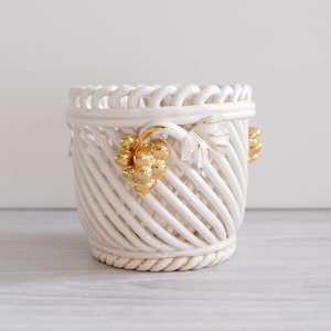 May include: A white ceramic basket-style planter with a woven design. The planter features gold-colored grape cluster accents and a rope-like trim. The interior is hollow, and the planter is suitable for small plants or decorative use.
