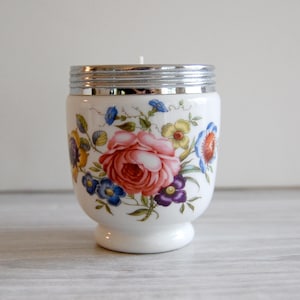 May include: A white ceramic candle holder with a rounded base and a metallic silver rim. The exterior features a colorful floral pattern with a large central pink rose surrounded by smaller blue, yellow, and purple flowers and green leaves.