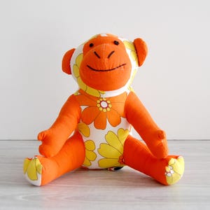 May include: A plush monkey toy with orange and yellow floral pattern fabric. The monkey has orange arms, legs, and ears, with a white face and a smiling expression. The toy is sitting on a light-colored surface.