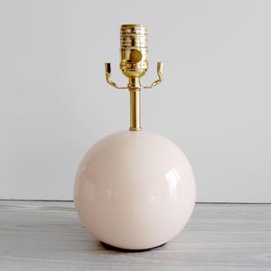 May include: A blush pink sphere-shaped table lamp with a brass-colored neck and bulb fixture. The lamp has a glossy finish and a simple, modern design. The lamp is approximately 12 inches tall.