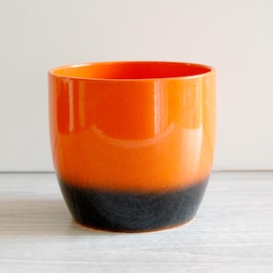 May include: A cylindrical ceramic planter featuring a smooth, glossy finish. The upper portion of the vessel is a vibrant, bright orange color, which transitions into a deep, solid black at the base, creating a distinct two-tone gradient effect.