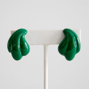May include: A pair of glossy, emerald green clip-on earrings. Each earring has a curved, organic shape with a smooth, reflective surface. The earrings are displayed on a white stand against a plain background, highlighting their vibrant color and design.