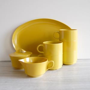 May include: A collection of bright yellow kitchenware, including a large oval platter, a sugar bowl with a lid, a creamer, and several stacked mugs. The set is arranged on a light-colored surface.