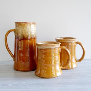 May include: Three handcrafted ceramic mugs and a pitcher in shades of brown and tan. The pitcher is tall with a large handle, while the mugs have a more traditional shape. The rims have a lighter, textured glaze.