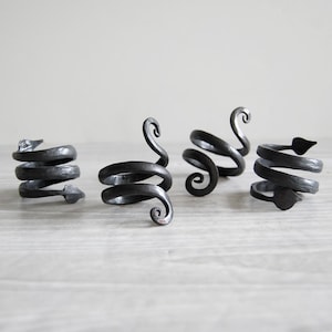 May include: Four handcrafted, dark gray metal napkin rings. Each ring features a unique, spiraling design with decorative elements such as an arrow, a curled end, and a leaf shape. The rings are displayed on a light gray surface.