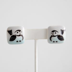 May include: A pair of square, white ceramic stud earrings. Each earring features a black and white cartoon bird design with a light blue background. The earrings are displayed on a white earring stand.