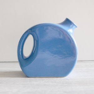 May include: A glossy, light blue ceramic pitcher with a rounded, circular body and a flat base. The pitcher features a side handle integrated into the circular frame and a small, angled spout at the top right, showcasing a smooth, reflective finish.
