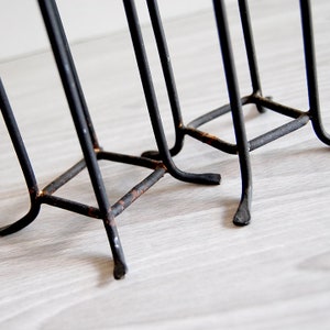 Puede incluir: Primer plano de la base de un revistero de metal negro. El revistero tiene tres patas y est&aacute; hecho de finas varillas de metal. El metal est&aacute; ligeramente oxidado.