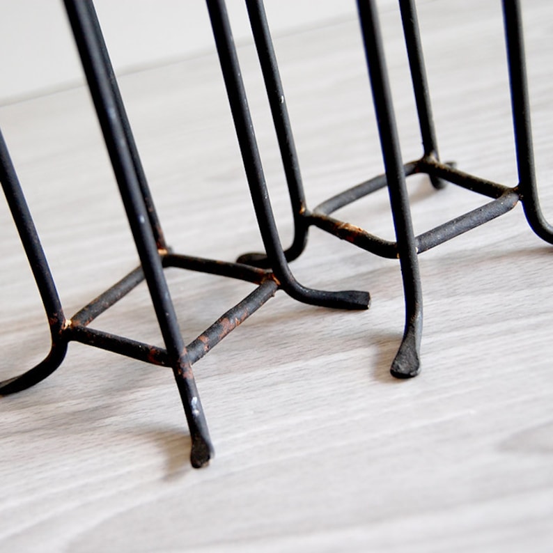 Puede incluir: Primer plano de la base de un revistero de metal negro. El revistero tiene tres patas y est&aacute; hecho de finas varillas de metal. El metal est&aacute; ligeramente oxidado.