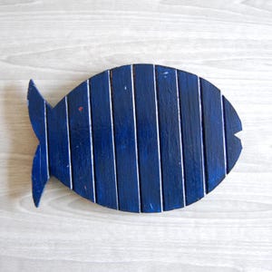 May include: A fish-shaped wall hanging constructed from vertical wooden slats painted in a deep navy blue. The object features a distinct tail fin on the left side and a small V-shaped notch on the right side, with visible wood grain texture.